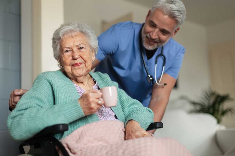 Resident gently holds a small dog in a bright assisted living apartment beside a window.